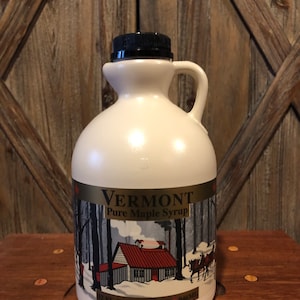 May include: A white plastic jug with a black lid and handle. The jug contains Vermont Pure Maple Syrup. The label features a winter scene with a red-roofed cabin, trees, and a horse-drawn sleigh. The label also includes the text "Vermont Pure Maple Syrup", "1 quart", "32 fl oz", and "946 ml".