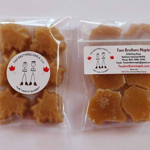 May include: Two clear plastic bags filled with maple candy. The candy is shaped like maple leaves. One bag has a sticker with the text "Two Brothers Maple LLC" and a cartoon of two brothers. The other bag has a sticker with the text "Two Brothers Maple" and contact information.