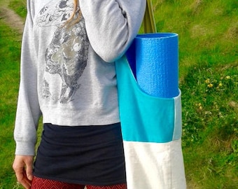 Puede incluir: Una mujer lleva una esterilla de yoga azul en una bolsa de lona multicolor con fondo blanco. La bolsa tiene una sección azul turquesa y una sección azul claro. La mujer lleva una sudadera gris con un estampado gráfico en blanco y negro, un cinturón negro y pantalones con estampado rojo y blanco.