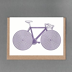Pode incluir: Um cartão com uma ilustração de bicicleta roxa. A bicicleta tem rodas com desenhos de círculos concêntricos. O cartão está em um fundo branco com uma borda marrom, em um fundo cinza.