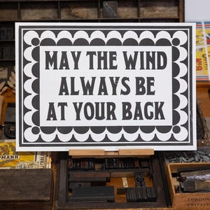 Op de afbeelding: Een zwart-wit bord met de tekst "MAY THE WIND ALWAYS BE AT YOUR BACK". Het bord heeft een decoratieve rand en wordt weergegeven op een houten drukpers.