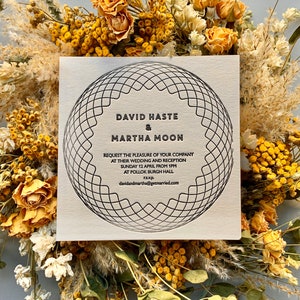 May include: A wedding invitation featuring the names "DAVID HASTE & MARTHA MOON" in black, enclosed within a geometric pattern. The card displays event details and is set against a backdrop of dried flowers and foliage.