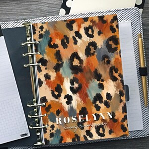 May include: A six-ring planner with a leopard print cover featuring the text ROSELYN twenty twenty-seven, containing a to-do list insert with the text TO DO LIST, a polka dot pen, a notes clipboard, and a small circular sticker with text.