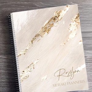 Może przedstawiać: Spiralny notatnik w kremowym kolorze z akcentami ze złotych liści. Okładka zawiera imię "Roselynn" i tekst "Meraki Planners" w złotym skrypcie. Ozdobna miska w kształcie ananasa zawiera kolorową taśmę washi.
