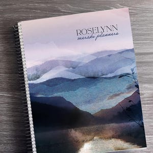 Puede incluir: Un planificador con espiral con un dise&ntilde;o de paisaje en acuarela. La portada presenta una cordillera en tonos azules y morados, con el texto "ROSELYNN meraki planners". Cinta washi decorativa y un arreglo floral son visibles.