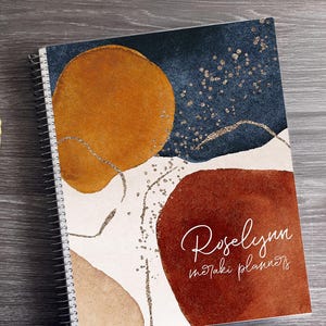 May include: Spiral-bound notebook with an abstract design featuring orange and brown shapes on a white background, with a navy blue top section. The notebook has the text "Roselynn motaki planners" in cursive.