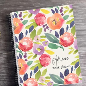 May include: A spiral-bound planner with a floral design. The cover features watercolor-style flowers in shades of red, orange, and purple, with green leaves on a white background. The name "Adrienne" and "meraki planners" are written on the cover.