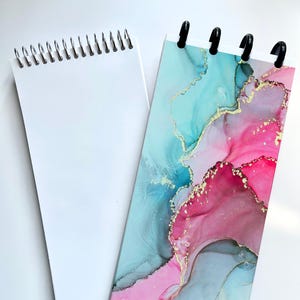 May include: Two notebooks. One has a white spiral binding and blank pages. The other has a colorful abstract design in shades of pink, blue, and gold. The second notebook has black plastic binding posts.