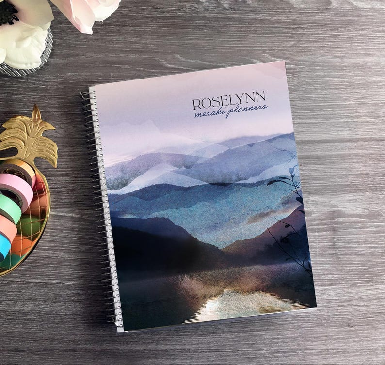 Puede incluir: Un planificador con espiral con un dise&ntilde;o de paisaje en acuarela. La portada presenta una cordillera en tonos azules y morados, con el texto "ROSELYNN meraki planners". Cinta washi decorativa y un arreglo floral son visibles.