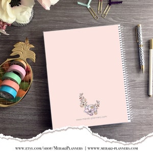 May include: A pink spiral-bound planner with a floral design and the website address meraki-planners.com. A gold pineapple-shaped dish holds colorful washi tape rolls. A pen with black polka dots and a white pen are also visible.
