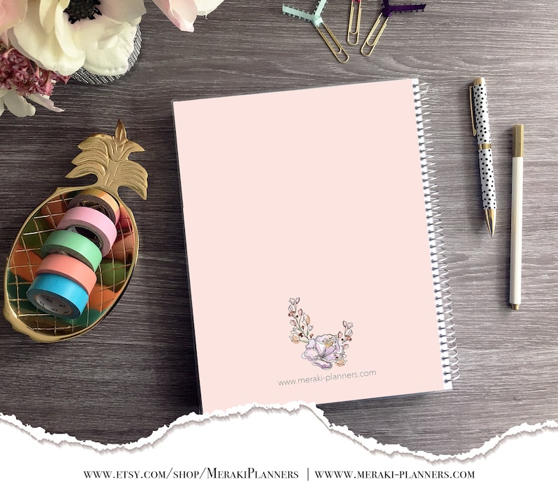 May include: A pink spiral-bound planner with a floral design and the website address meraki-planners.com. A gold pineapple-shaped dish holds colorful washi tape rolls. A pen with black polka dots and a white pen are also visible.