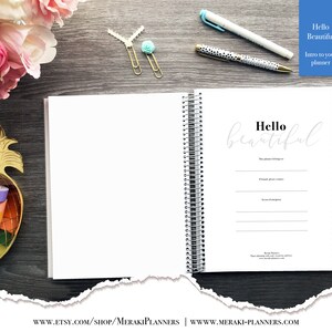 May include: Open spiral-bound planner with the words "Hello Beautiful" on the inside page. The planner is white with black and white striped edges. A gold pineapple-shaped container holds colorful washi tape.