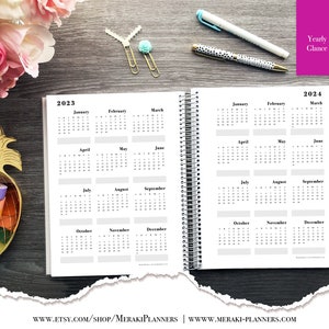 May include: Open spiral-bound planner displaying yearly calendars for 2023 and 2024. The pages are white with black text, showing months and dates. The planner is open on a dark wood surface, with pens and decorative items nearby.