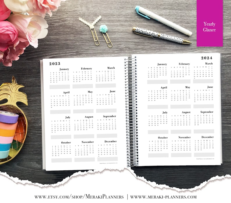 May include: Open spiral-bound planner displaying yearly calendars for 2023 and 2024. The pages are white with black text, showing months and dates. The planner is open on a dark wood surface, with pens and decorative items nearby.