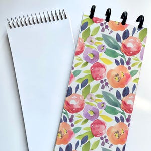 May include: Two spiral-bound notepads. One notepad has a blank white cover and pages. The other notepad has a floral design with red, orange, and purple flowers, green leaves, and black binding clips.