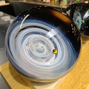 May include: A clear glass sphere with a swirling galaxy design in shades of white, blue, and black. A small, colorful orb is suspended within the center. The sphere is displayed on a wooden surface.