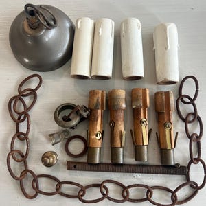 May include: An assortment of vintage chandelier parts, including a metal ceiling canopy, four off-white candle covers, four wooden and brass bulb holders, a rusty chain, and various hardware pieces. The components are arranged on a light-colored surface.