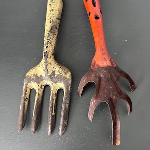 May include: Two vintage garden hand tools. One is a yellow and brown metal fork with four tines. The other is a red and brown metal tool with a leaf-like design and holes in the handle. Both tools show signs of wear.