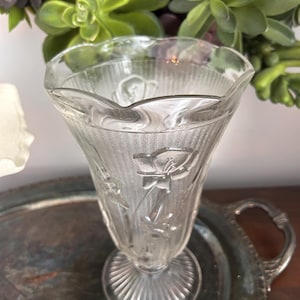 May include: A clear glass vase with a fluted design and floral pattern. The vase has a wide mouth and a narrow base.