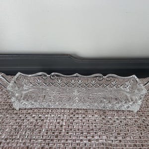 May include: A clear, rectangular glass serving dish with ornate detailing. The dish features a diamond pattern and curved handles on each end. The edges have a decorative, scalloped design. The dish is empty and sits on a textured surface.