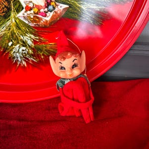 May include: A vintage Christmas elf ornament. The elf has a red hat and outfit, with a green and silver collar. The elf has a smiling face with painted features. The ornament is on a red surface.
