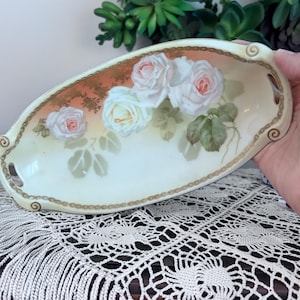 May include: Oval porcelain dish with a floral rose design. The dish features a cream-colored base with a gold trim and handles. The roses are painted in shades of pink and white with green leaves.