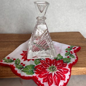 May include: A clear glass perfume bottle with a square base and a geometric design. The bottle is sitting on a white handkerchief with a red poinsettia design.