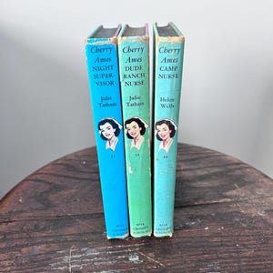 May include: Three vintage Cherry Ames novels, featuring titles like "Night Supervisor" and "Dude Ranch Nurse." The books have colorful spines in blue, green, and teal, with illustrations of a woman in a nurse's cap. The books are displayed on a wooden surface.