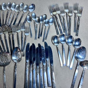 May include: A collection of vintage silverware, including forks, spoons, knives, and serving utensils. The silver-toned flatware is arranged on a neutral surface, showcasing various designs and sizes. The set includes multiple forks, spoons, and knives, ideal for a formal dining setting.