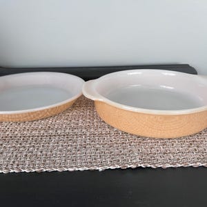 May include: Two vintage oval and round baking dishes. The dishes have a light peach exterior with a textured pattern and white interiors. One dish has handles. They are placed on a woven placemat.