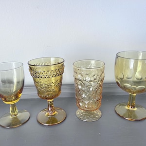 May include: Four amber-colored vintage drinking glasses. The glasses vary in shape and design, including a stemmed wine glass, a goblet with a textured pattern, a short tumbler, and a large stemmed glass with a rounded bowl. The glasses are arranged on a gray surface.