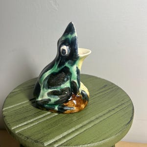 May include: A ceramic frog-shaped pitcher with a glossy finish, featuring a color palette of deep black, vibrant green, and earthy brown. The frog is depicted in a seated position with its mouth open, serving as the spout for the small vessel.