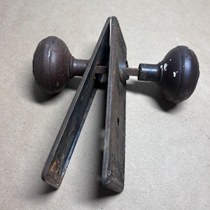 May include: An antique door knob set with two round knobs and a rectangular backplate. The knobs and backplate are made of metal with a rusty, aged appearance. The set includes a keyhole.