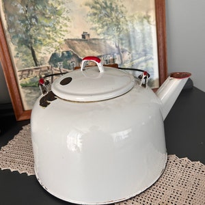 May include: White enamel teapot with a red handle and spout. The teapot has a black rim and a black spot on the side. The teapot is sitting on a crocheted doily.