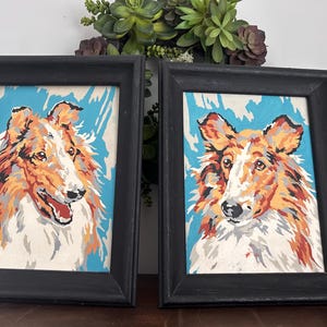 May include: Two framed paintings of Collie dogs. The paintings are done in a paint-by-number style with a blue background. The frames are black.