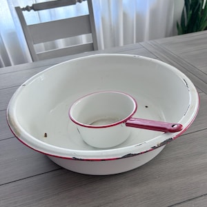 May include: A white enamelware wash basin with a red trim and a matching small pitcher. The basin has a distressed look, with chipped paint around the rim. The pitcher has a red handle. The items are on a gray wooden table.