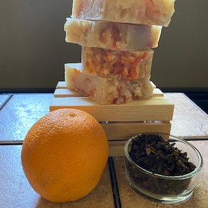 May include: Four bars of handmade soap stacked on a wooden crate. The soap is a light beige colour with orange flecks. An orange and a small bowl of dried cloves are in the foreground.
