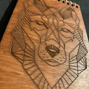 May include: A wooden notebook with a geometric wolf design engraved on the front cover. The notebook has a spiral binding and is made of natural wood.
