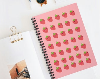 May include: A spiral-bound notebook with a pink cover featuring a pattern of smiling cartoon strawberries. The notebook is open, revealing blank white pages. Sunglasses and a gold binder clip are also visible.