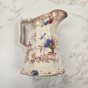 Op de afbeelding: Crèmekleurige keramische kan met een handvat en een uitlopende basis. De kan is versierd met bloemen- en architecturale patronen in rood, blauw en geel. De rand heeft een bruine rand.