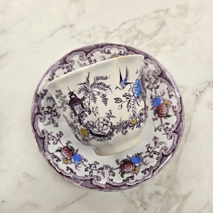Puede incluir: Un juego de taza y platillo de té vintage. La taza blanca presenta un diseño floral y de pagoda detallado con detalles en azul, amarillo y rojo. El platillo tiene un borde floral morado a juego. Un clásico.