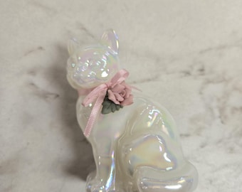 May include: A sitting cat figurine crafted from iridescent white glass with a glossy finish. The cat is adorned with a light pink satin ribbon tied around its neck, featuring a small, sculpted pink rose with two green leaves attached to the ribbon.