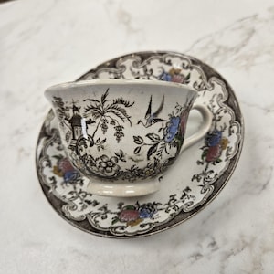 May include: A vintage teacup and saucer set. The cup and saucer are white with a brown and black floral and landscape design. The cup has a handle and sits on a matching saucer. The set is likely made of porcelain.