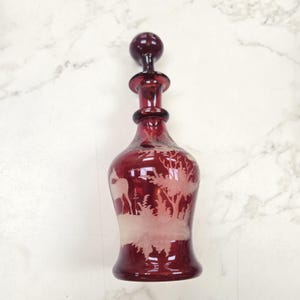 May include: A red glass decanter with a clear glass stopper. The decanter is etched with a white design of a deer in a forest.