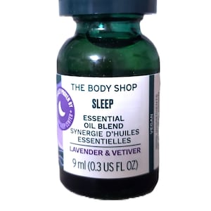 Könnte beinhalten: Eine kleine grüne Glasflasche mit einem weißen Etikett, auf dem "THE BODY SHOP SLEEP ESSENTIAL OIL BLEND SYNERGIE D'HUILES ESSENTIELLES LAVENDER & VETIVER 9 ml (0.3 US FL OZ)" und ein violettes Mondlogo mit dem Text "PROVEN BY SLEEP CENTER" steht. Die Flasche ist mit "VEGAN" und "100% INGREDIENTS" gekennzeichnet.