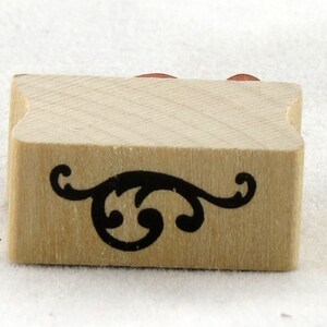 Puede incluir: Un sello de madera con un diseño decorativo negro en forma de espiral. El sello es rectangular con bordes redondeados.