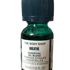 Puede incluir: Una pequeña botella de vidrio verde con una tapa verde oscuro. La botella contiene una mezcla de aceites esenciales para aromaterapia. La etiqueta dice "The Body Shop Breathe Essential Oil Blend Synergie d'Huiles Essentielles Eucalyptus & Rosemary 9 ml (0.3 US FL OZ) Vegan".