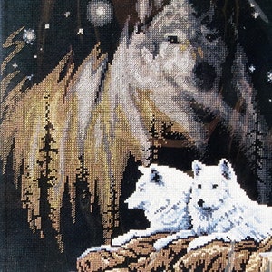 May include: A cross-stitch pattern depicting a white wolf with two smaller white wolves in the foreground. The wolves are set against a dark background with a starry sky and a forest.