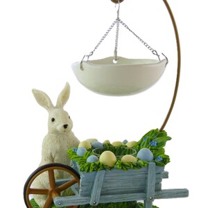 May include: An Easter-themed decorative piece featuring a white bunny next to a blue cart filled with eggs. A hanging white bowl is suspended above the cart. The base is green with grass and small blue flowers.