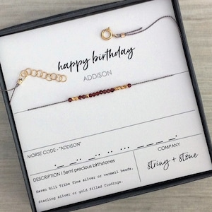 May include: A gray string bracelet with gold and burgundy beads. The bracelet is in a box with the words "happy birthday" and "ADDISON" written on it. The box also includes the company name "String + Stone" and a description of the bracelet.